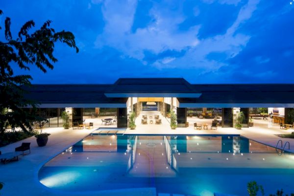 A luxurious building with a large, illuminated swimming pool at night, surrounded by lounge chairs, and trees in the foreground.