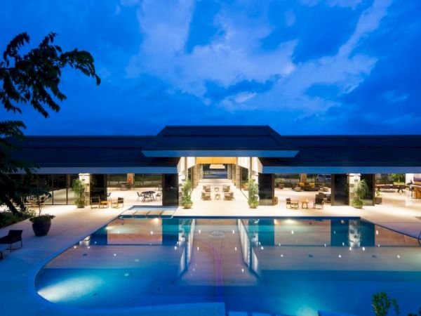 A luxurious building with a large, illuminated swimming pool at night, surrounded by lounge chairs, and trees in the foreground.