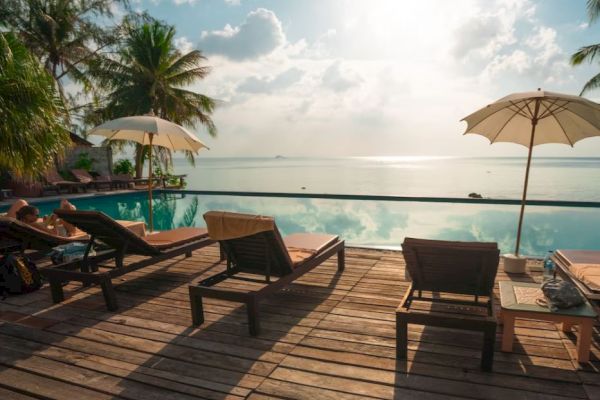 A tranquil seaside setting with sun loungers, umbrellas, palm trees, and an infinity pool overlooking the calm ocean waters under a bright sky.