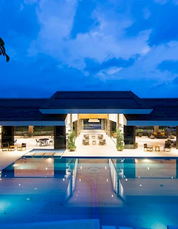 An outdoor pool area at night with a large, modern building in the background adorned with lights and lounge furniture.