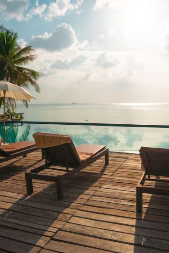 A serene poolside view with lounge chairs, umbrellas, and palm trees overlooking a calm sea under a partly cloudy sky with sunlight glistening.