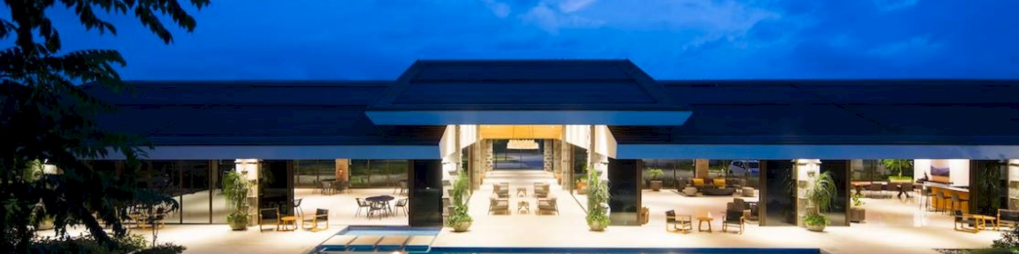 A modern, illuminated building with large glass windows and a spacious area features a central swimming pool with lounge chairs, pictured at dusk.