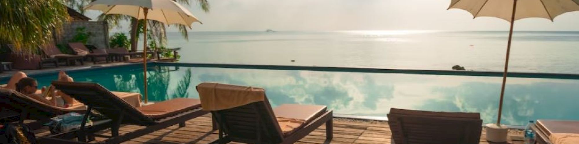 A serene poolside deck with lounge chairs and umbrellas overlooking a calm ocean under a partly cloudy sky during sunrise or sunset.