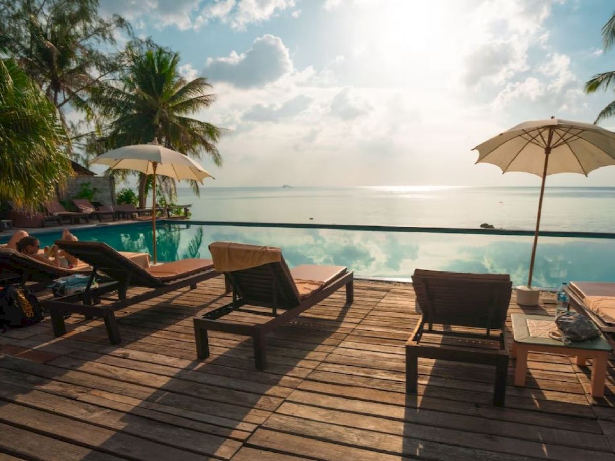A serene poolside scene with lounge chairs, umbrellas, and a stunning view of the ocean and sky, framed by palm trees on a sunny day.