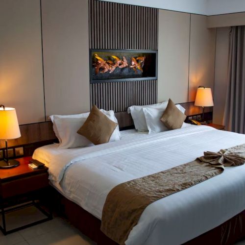 A modern hotel room with a large bed, bedside tables with lamps, neutral tones, a wall-mounted decorative panel, and a window with curtains.