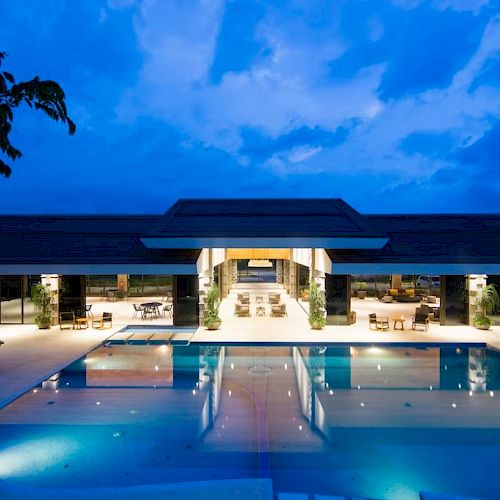 The image shows a luxurious modern building with extensive open spaces and a large, illuminated swimming pool in the foreground at twilight.