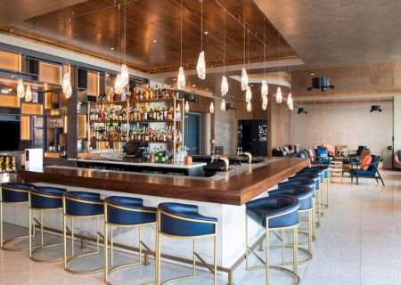 A modern bar with blue cushioned stools, a variety of liquor bottles behind the counter, and elegant lighting fixtures.
