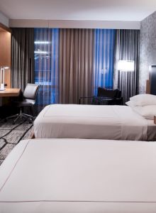 This image features a modern hotel room with two neatly made beds, a wall-mounted TV, a desk with a chair, and contemporary lighting fixtures.