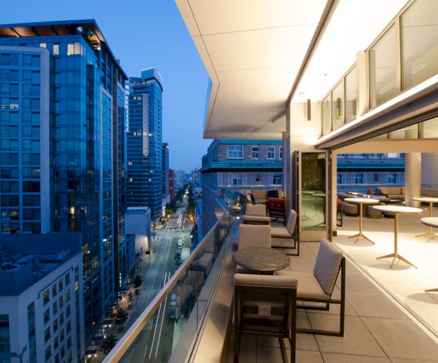 The image shows a modern cityscape at dusk with high-rise buildings and a balcony equipped with tables and chairs, offering a scenic view.