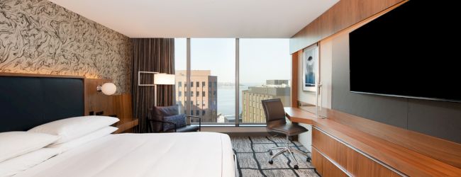A modern hotel room with a large bed, wall-mounted TV, desk, chair, and a view of buildings and water through a large window.