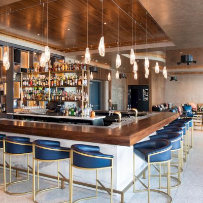 The image showcases a modern bar with a wooden counter, blue bar stools, hanging lights, and a well-stocked liquor shelf, situated in a spacious, stylish lounge.