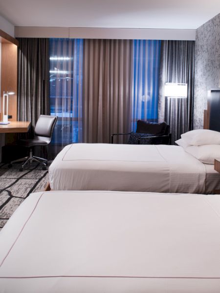 A modern hotel room features two neatly made beds, a mounted TV, a work desk with a chair, and contemporary decor with patterned walls and floor.