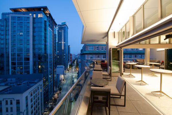 This image shows a modern rooftop terrace with furniture overlooking a city street and high-rise buildings at dusk, creating a stylish urban setting.