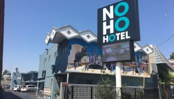 The image shows a building labeled "NoHo Hotel" with a colorful mural, a few cars parked nearby, and a clear, sunny sky.