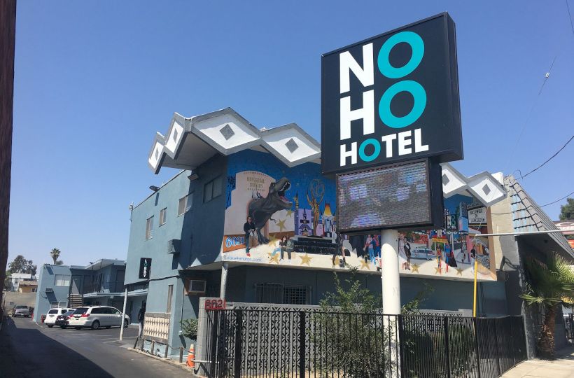 The image shows a building labeled "NoHo Hotel" with a colorful mural, a few cars parked nearby, and a clear, sunny sky.