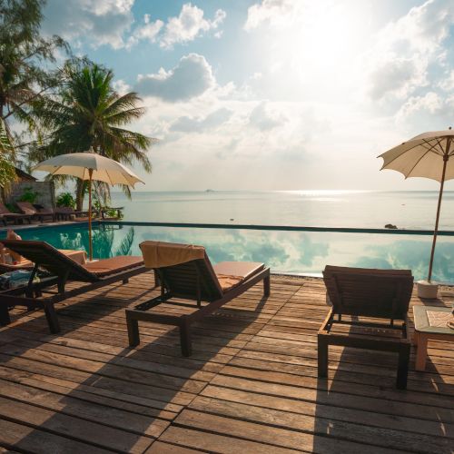 Sunlit poolside with lounge chairs, umbrellas, and a serene ocean view, surrounded by tropical trees, creating a relaxing and scenic atmosphere.
