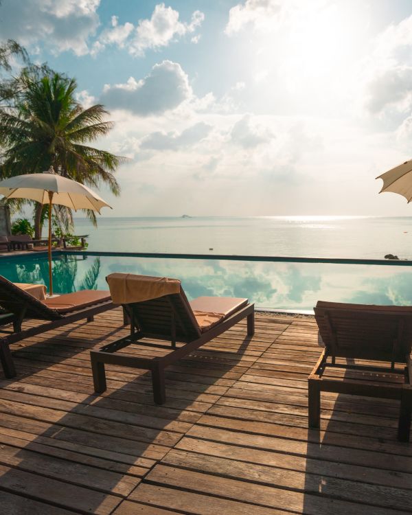 Sunlit poolside with lounge chairs, umbrellas, and a serene ocean view, surrounded by tropical trees, creating a relaxing and scenic atmosphere.