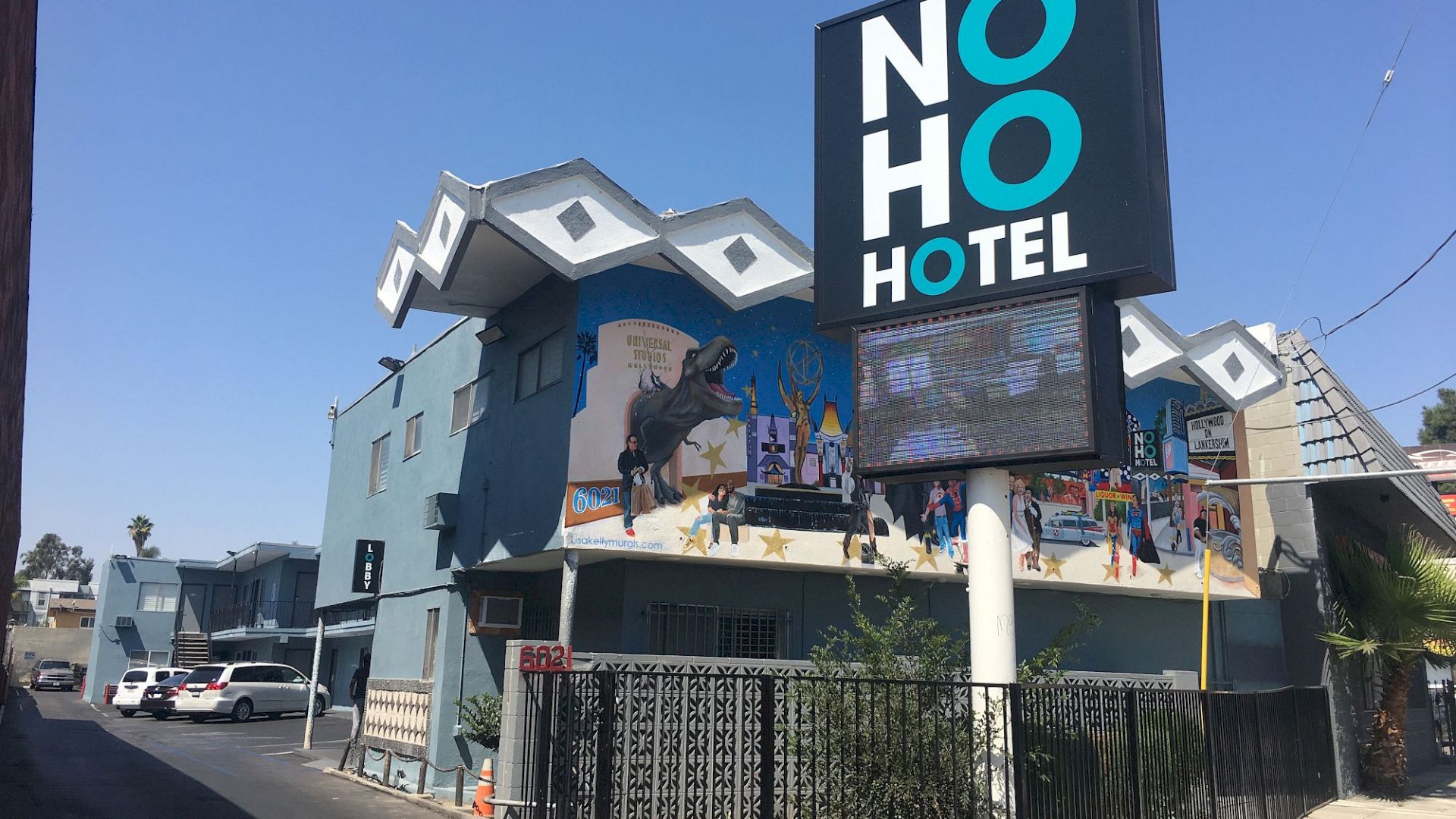 The image shows a blue NoHo Hotel building with a sign at the forefront. The building features a mural and a unique roof design.