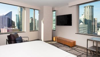 A modern hotel room with large windows displaying a city skyline, a wall-mounted TV, a bed, chair, and a small table near the window ending the sentence.