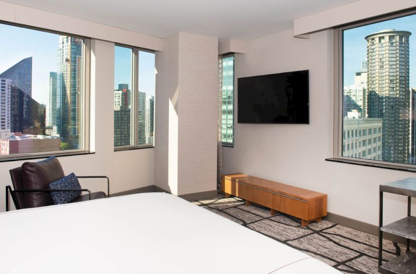 A modern hotel room with large windows displaying a city skyline, a wall-mounted TV, a bed, chair, and a small table near the window ending the sentence.