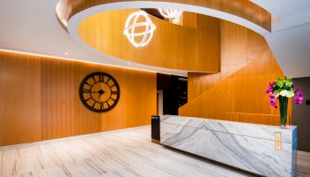 A modern lobby features a marble reception desk, wooden paneling, a large wall clock, decorative lighting, and a vase with flowers on the counter.