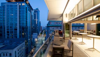 A cityscape view from a modern balcony at dusk, featuring outdoor seating, high-rise buildings, and a well-illuminated interior space.