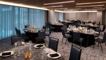 The image shows an elegantly arranged banquet hall with round tables set for a formal event, featuring black chairs, white flowers, and dim lighting.