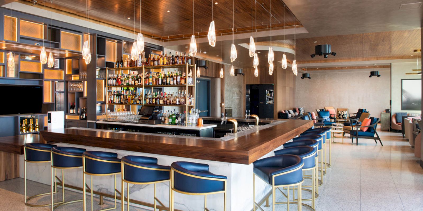 A modern bar with blue stools, a stocked liquor shelf, pendant lights, and a seating area in the background.