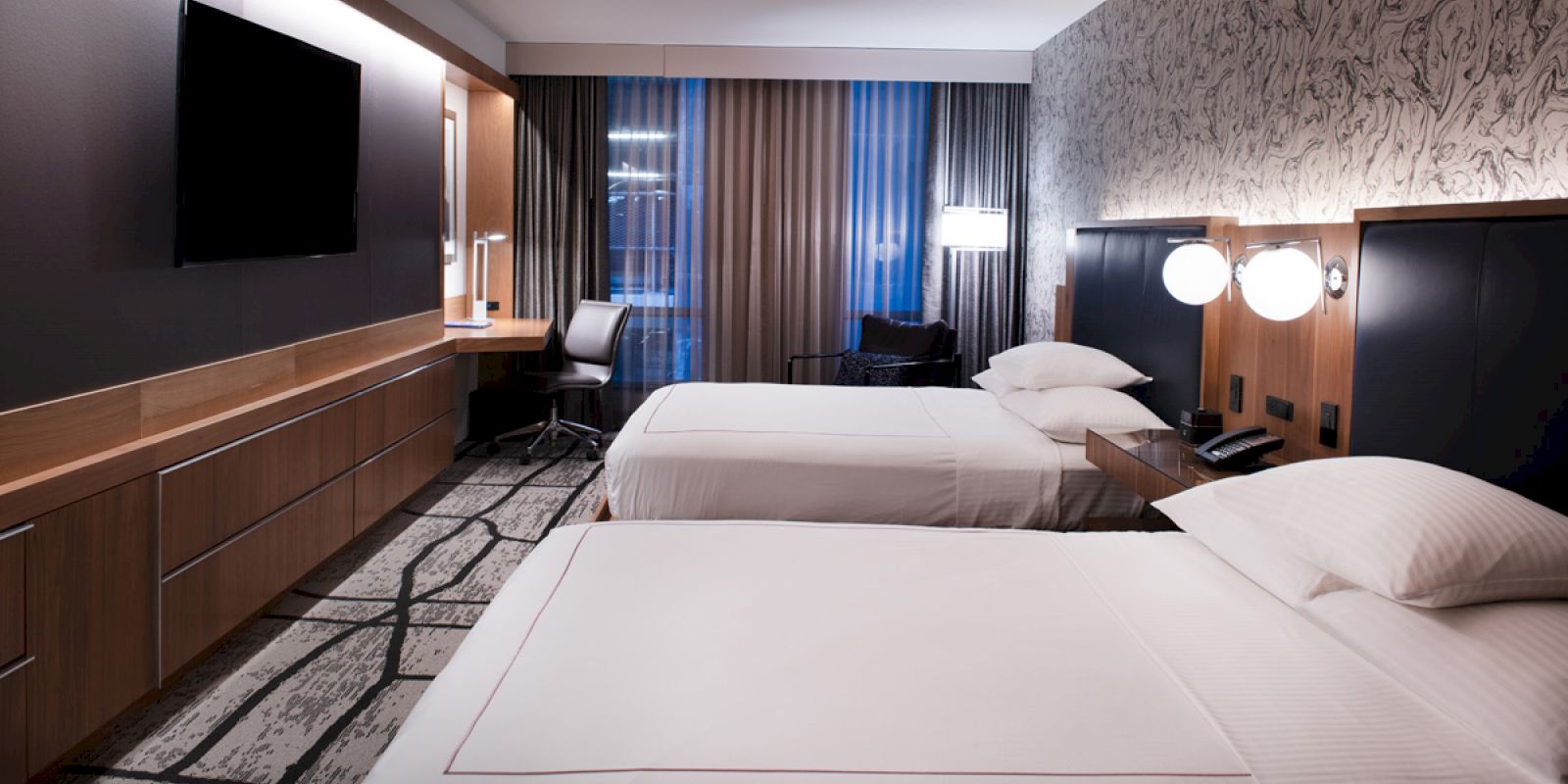 This image shows a modern hotel room with two beds, a flat-screen TV, a desk with a chair, and large windows with curtains.