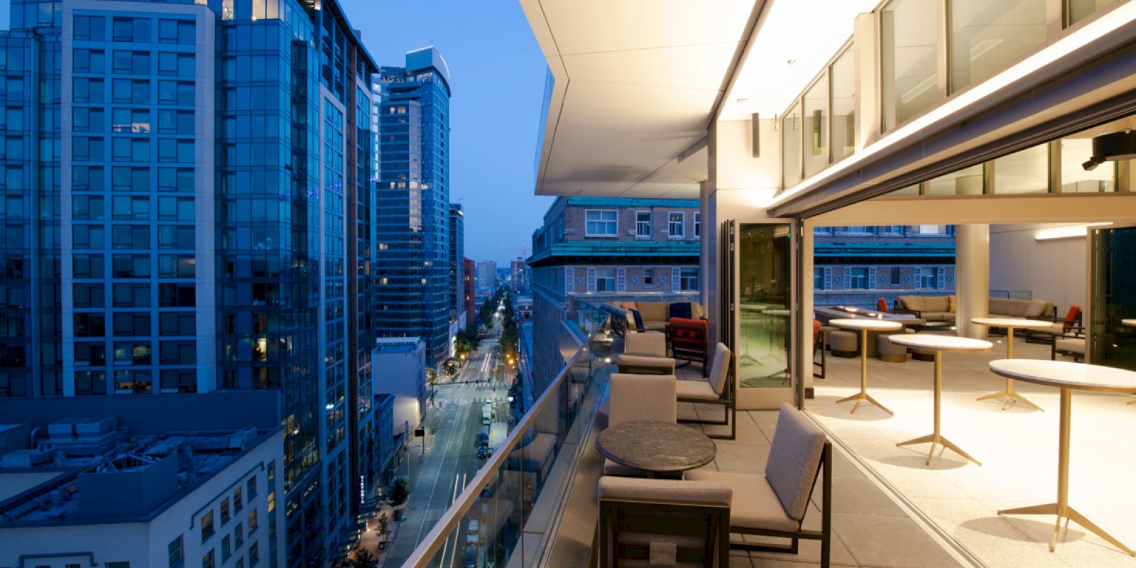 A modern urban balcony with seating and tables overlooks a city street at twilight, surrounded by tall buildings with large windows.