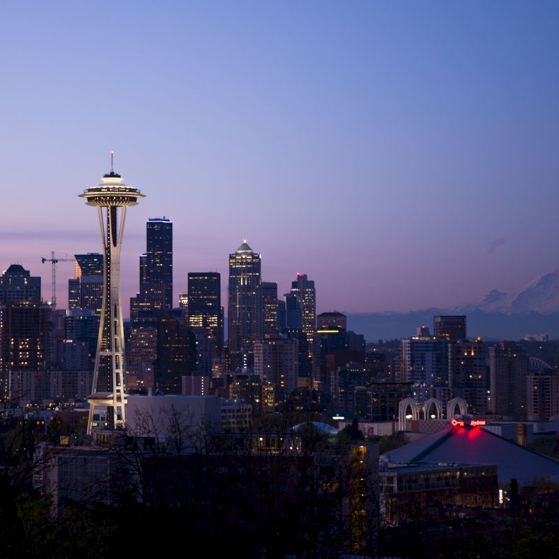 The image shows a nighttime cityscape with the Space Needle prominently visible, surrounded by tall buildings, and Mt. Rainier in the background.