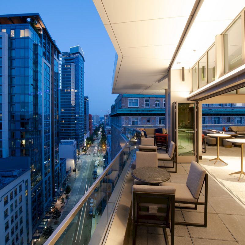 A modern balcony scene overlooking an urban street at dusk, featuring outdoor seating and tables with high-rise buildings in the background.