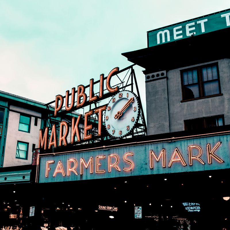 The image shows the iconic neon signs for "Public Market" and "Farmers Market" with a clock at a famous outdoor market.
