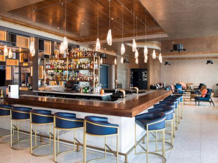 A modern bar with a sleek wooden counter, numerous high chairs, hanging pendant lights, and a fully stocked wall of beverages in a spacious, stylish room.