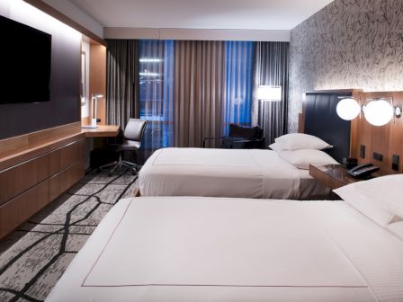 The image shows a modern hotel room with two beds, a wall-mounted TV, a desk with a chair, ambient lighting, and neutral decor.