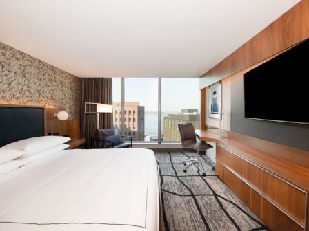 A modern hotel room features a large bed, desk, chair, extensive cabinetry, large TV, and a window with a city view.