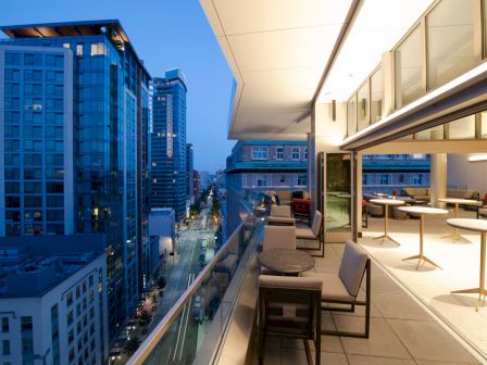 A modern rooftop terrace at sunset, featuring outdoor seating and tables, overlooking a vibrant cityscape with tall buildings and lit streets.