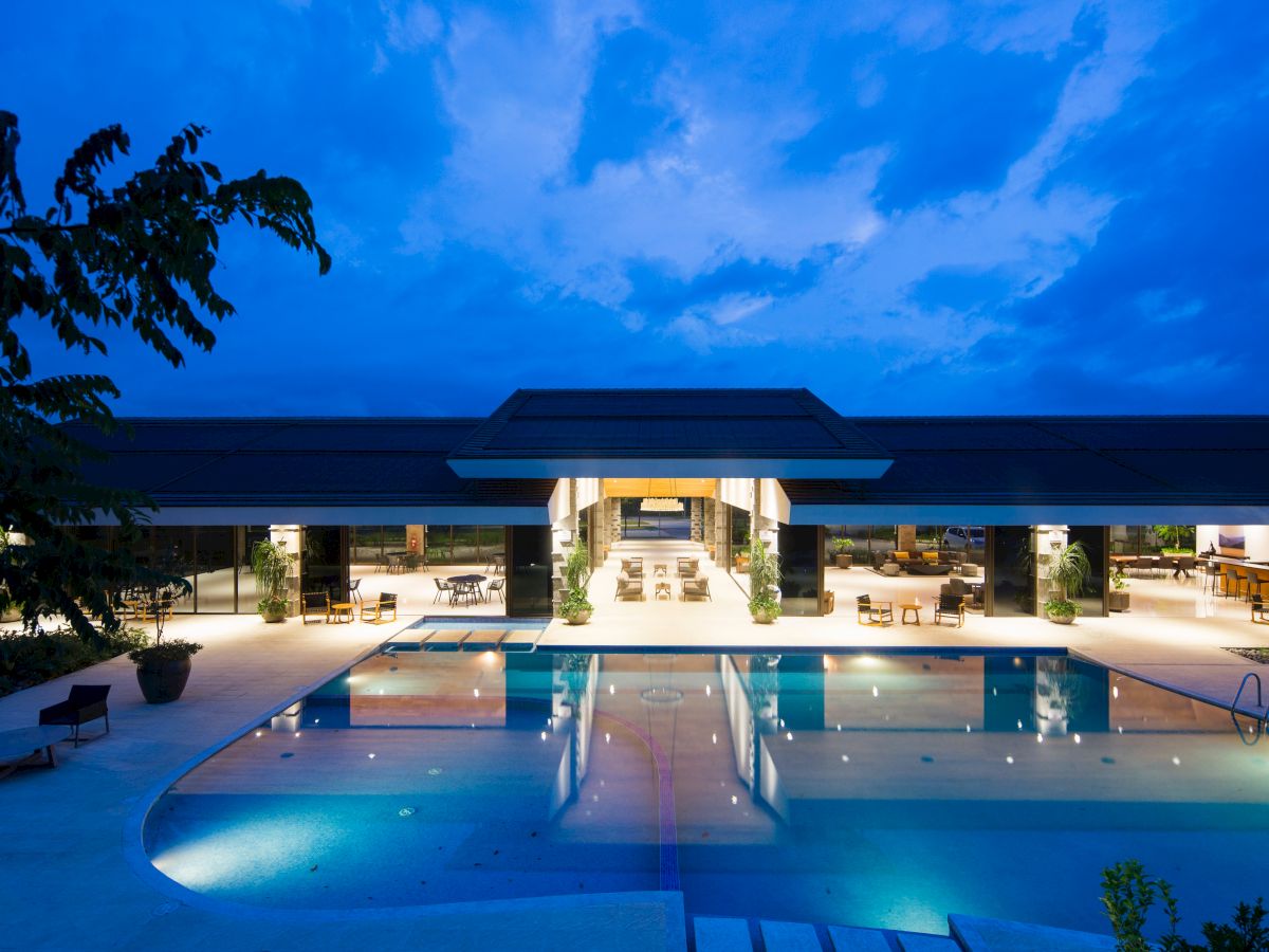 The image showcases a luxurious outdoor swimming pool area of a modern building at dusk, with bright interior lighting illuminating the space.