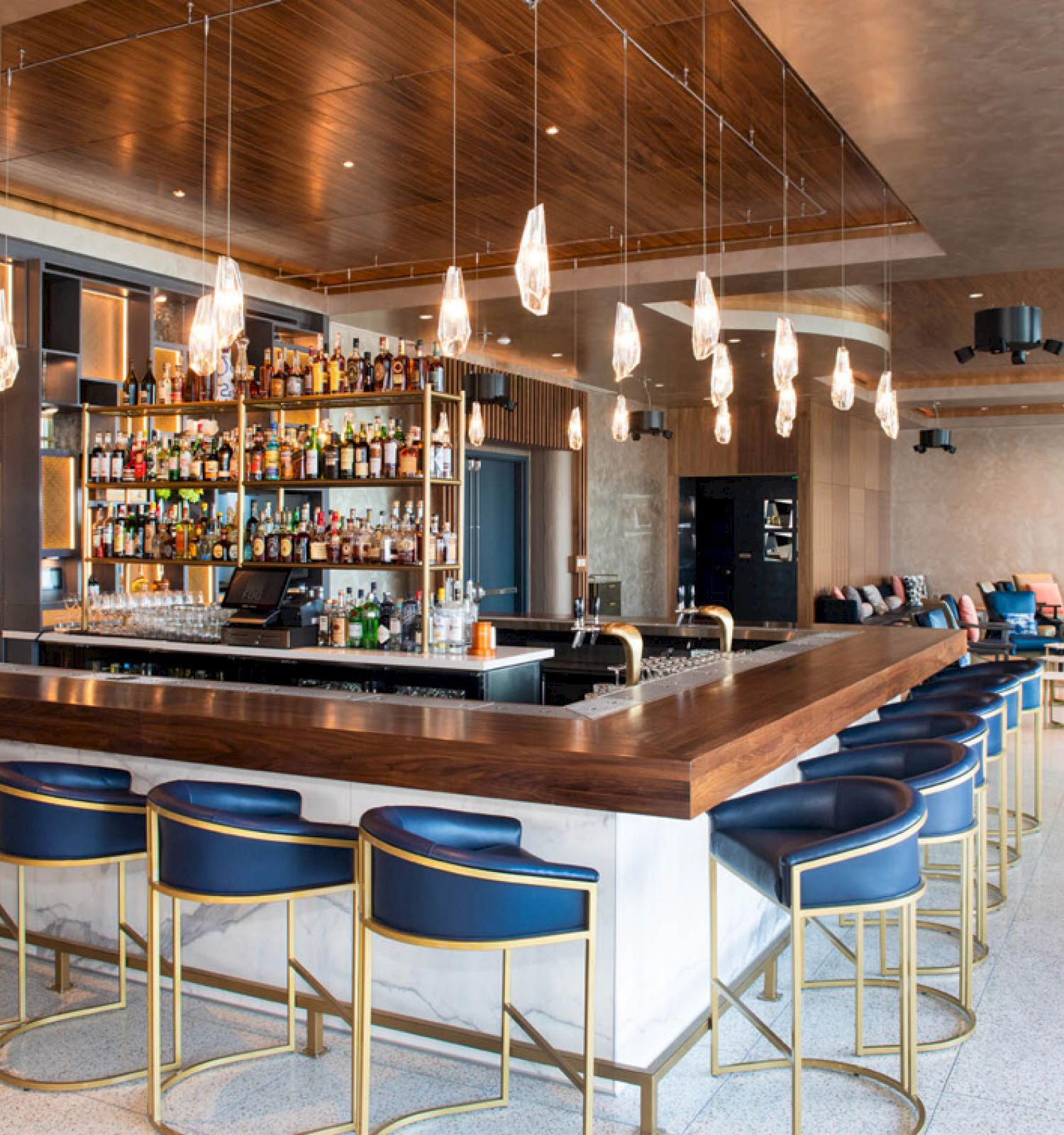 A modern bar with blue upholstered stools, overhead pendant lights, and a fully stocked liquor shelf behind the counter.