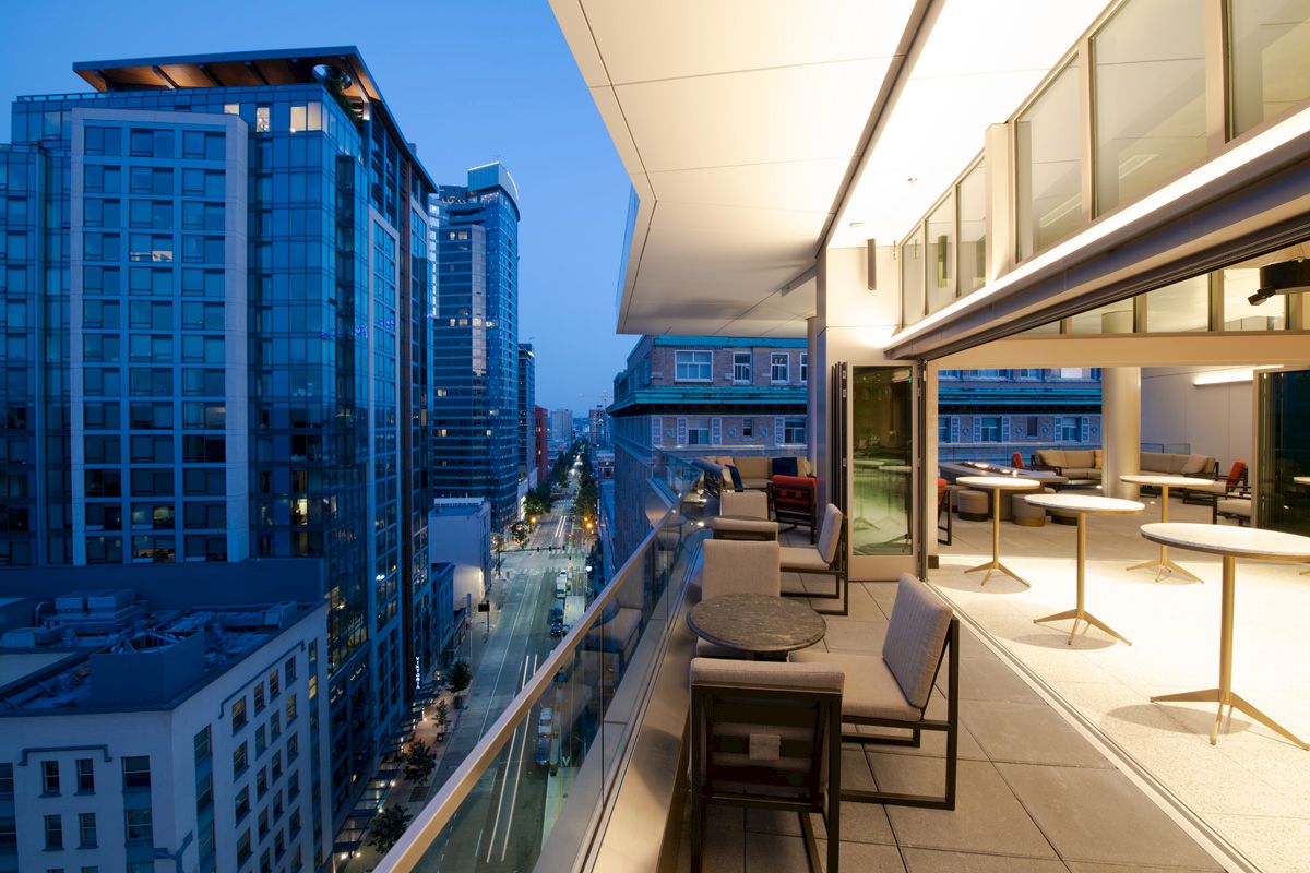 A modern cityscape at dusk with tall buildings and an outdoor patio featuring tables and chairs, overlooking a street below.