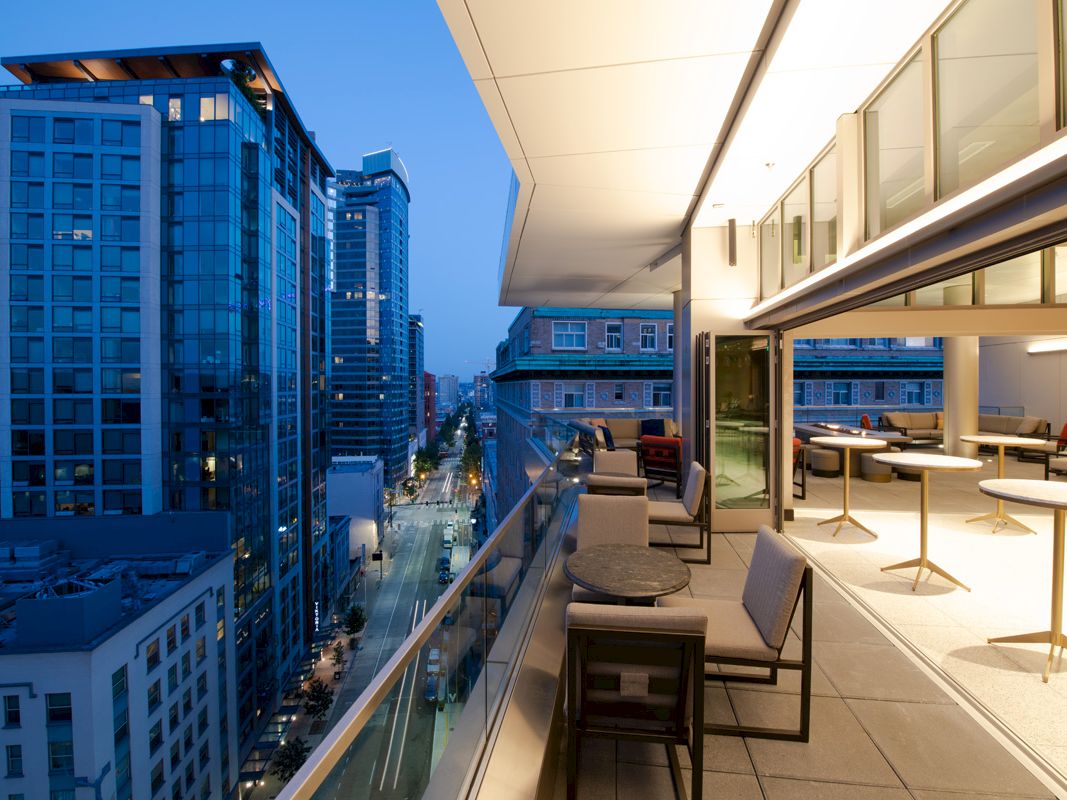 A modern cityscape at dusk with tall buildings and an outdoor patio featuring tables and chairs, overlooking a street below.