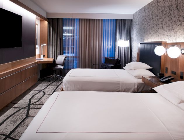 A modern hotel room with two beds, a desk with a chair, a wall-mounted TV, stylish decor, and large windows with curtains.