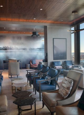 A modern lounge with numerous chairs and tables, large windows offering a scenic view of water and a Ferris wheel, and a cityscape mural on one wall.