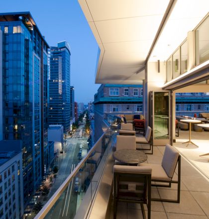 A modern urban balcony with a lounge area overlooks a cityscape of tall buildings lit up in the evening, creating an inviting atmosphere.