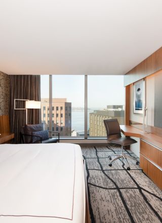A modern hotel room features a large bed, a desk with a chair, a flat-screen TV, and a city view through large windows.