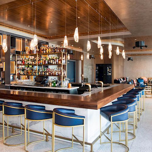 The image shows a modern bar with a wooden countertop and blue barstools around it. The bar has a variety of bottles and hanging lights above.