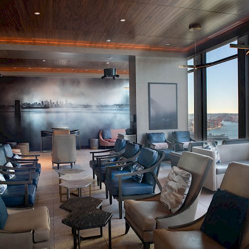 A modern lounge area with stylish chairs, panoramic windows showing a waterfront view, and a wall mural of a cityscape.