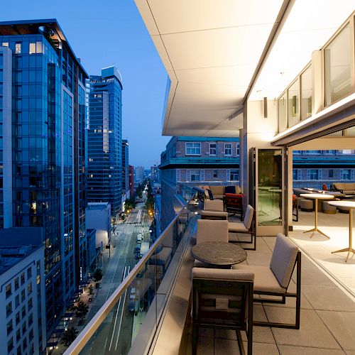A modern balcony with tables and chairs, overlooking a cityscape with tall buildings and a street below during evening time.