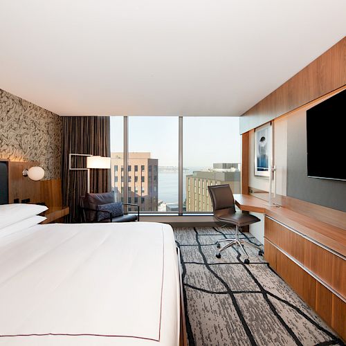 A modern hotel room with a large bed, wall-mounted flat-screen TV, desk with chair, floor-to-ceiling windows offering a city view ends the sentence.