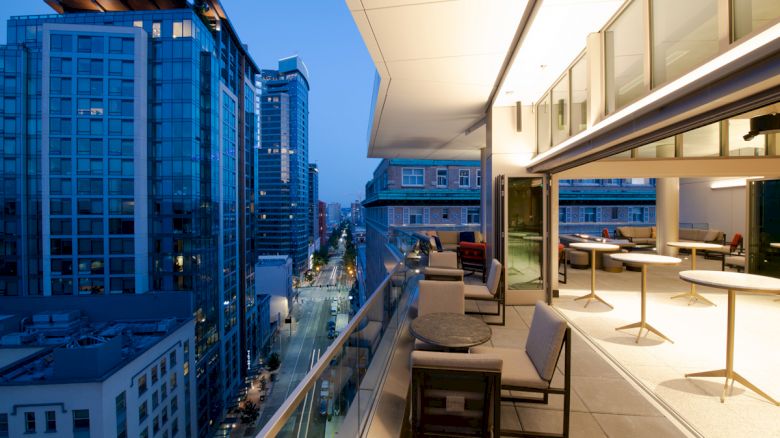 The image shows a modern balcony with seating overlooking a city street, surrounded by high-rise buildings during the evening.